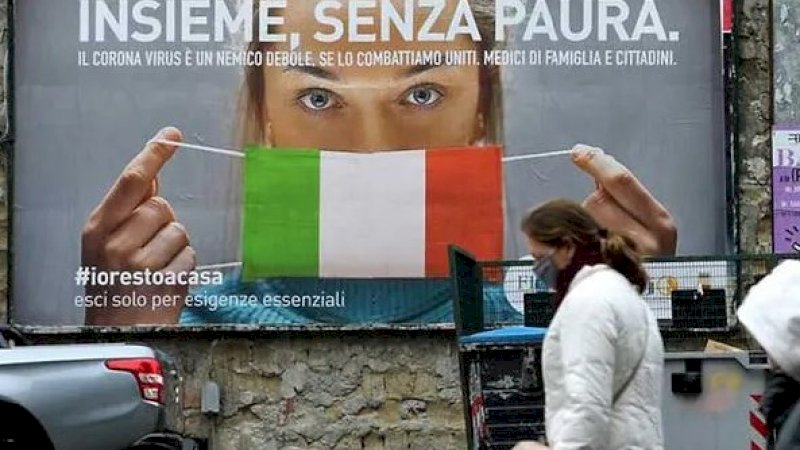 Orang-orang berjalan melewati papan iklan yang memperlihatkan seorang wanita mengenakan masker berwarna bendera Italia. (AFP)